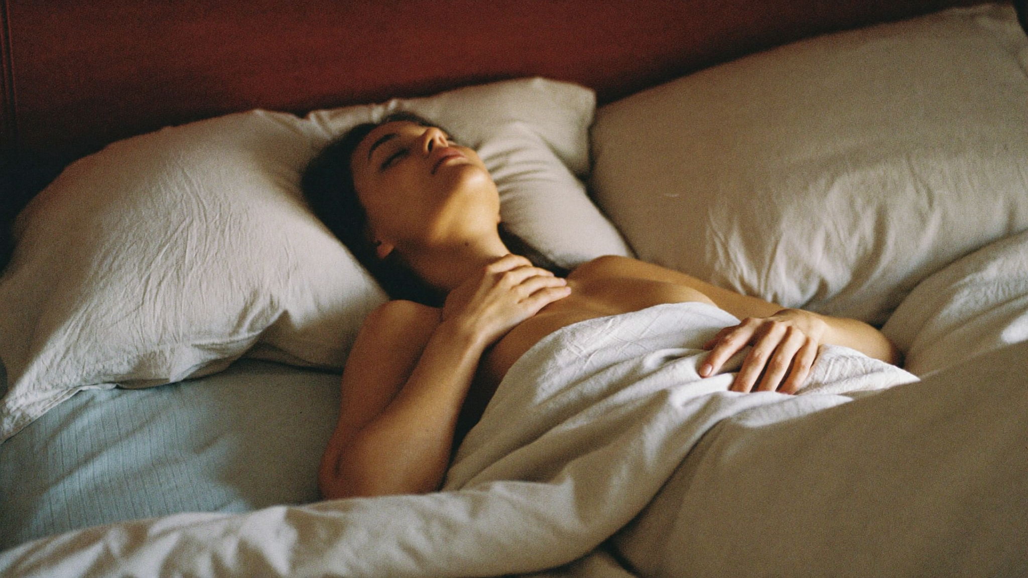 A woman resting on warm linen bedding in soft natural light, mid-exhale