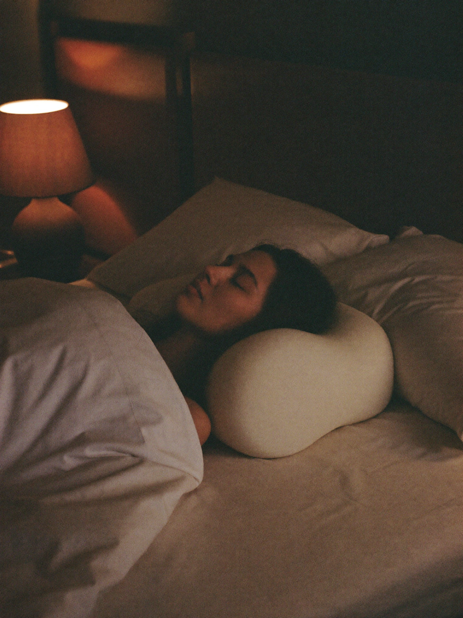 A woman at rest in bed on a curved cervical pillow in warm lamp light